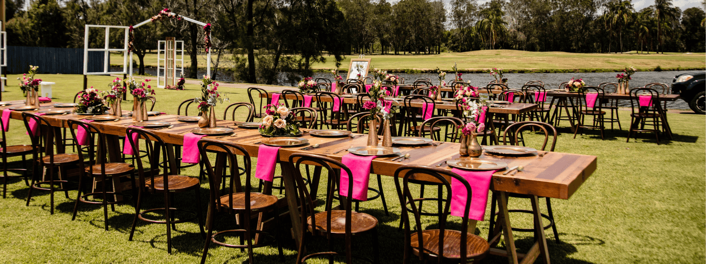 Long wooden tables with pink chairs and floral arrangements set on a grassy area by a lake.