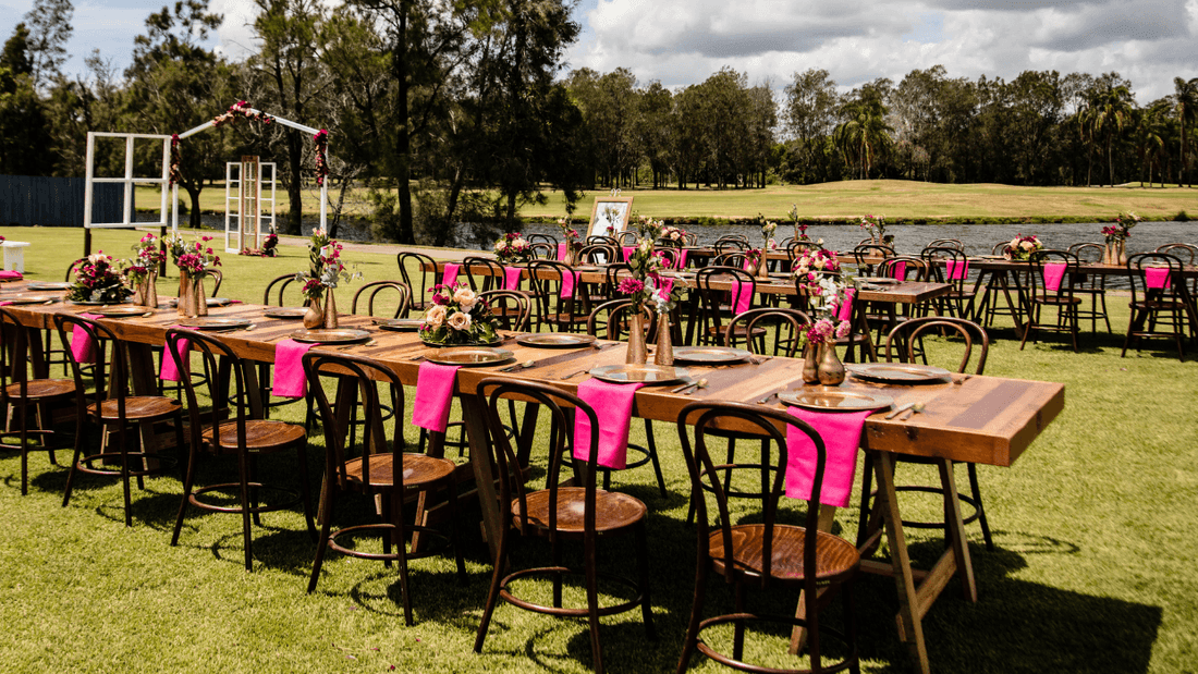 Long wooden tables with pink chairs and floral arrangements set on a grassy area by a lake.