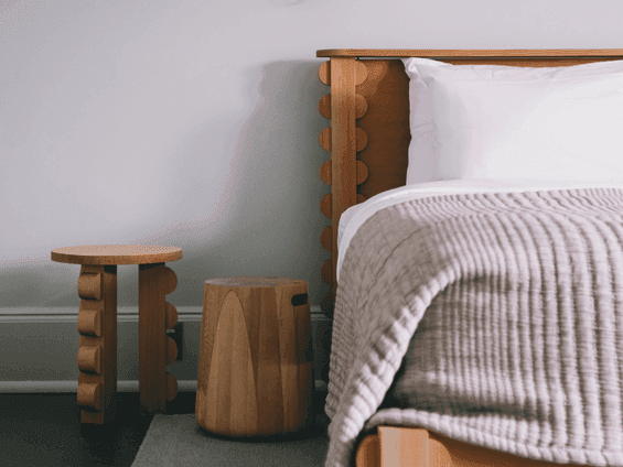Wooden bed with a white pillow and gray blanket, next to two wooden stools on a gray carpet.