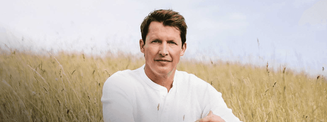 James Blunt sits outdoors among tall grass, with a serene landscape backdrop near Novotel Sydney Olympic Park