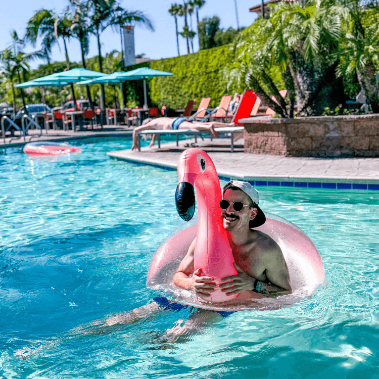 Guest relaxing in a pink flamingo pool float on a sunny day with palm trees and lounge chairs in the background.
