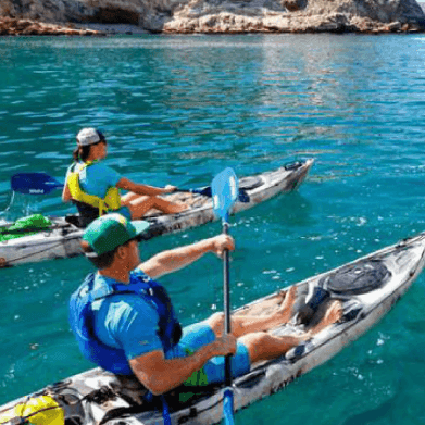 Two people kayaking on clear blue water with rocky shoreline in background.