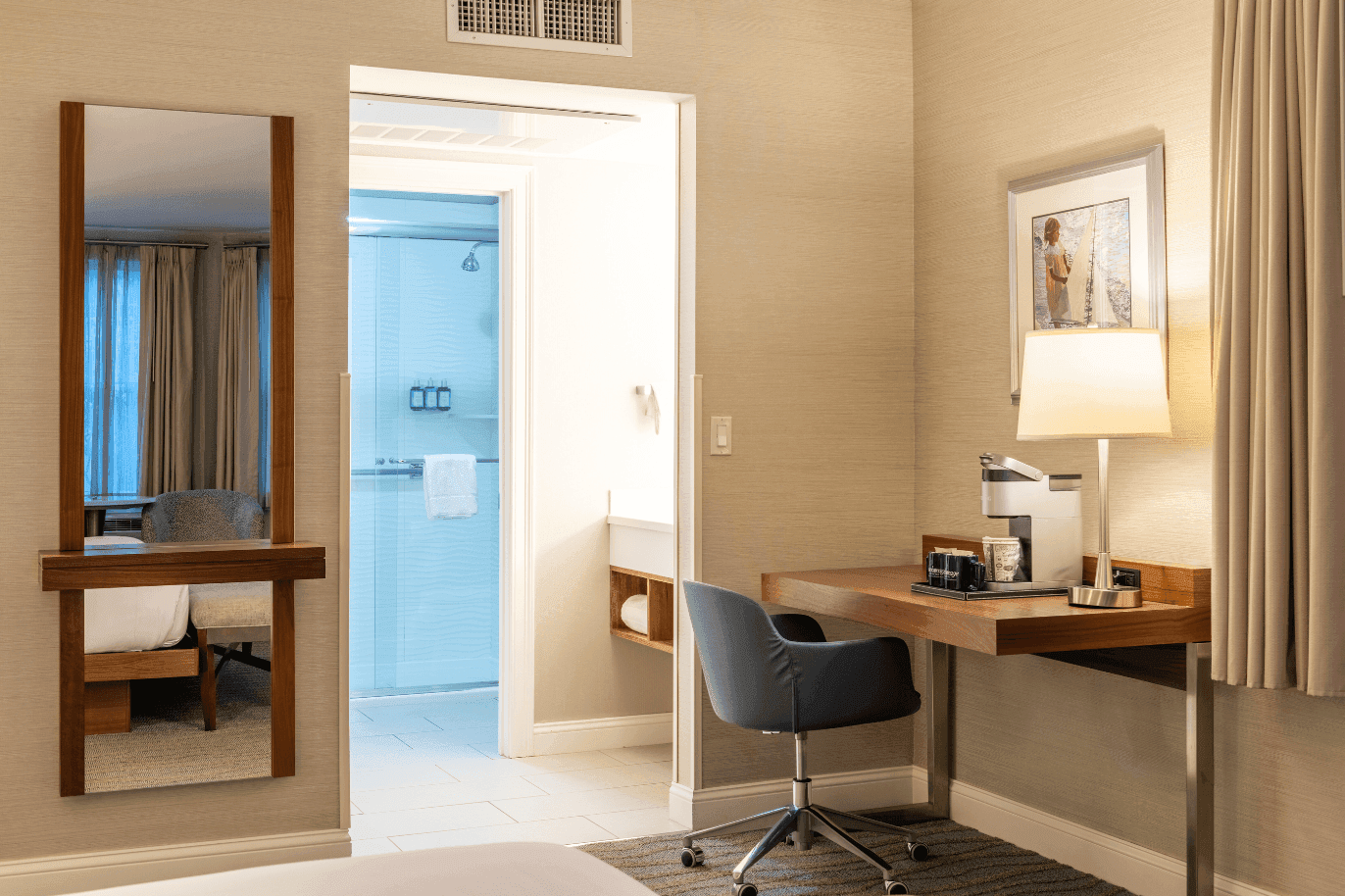A hotel room with a desk, chair, lamp, mirror, and doorway to bathroom