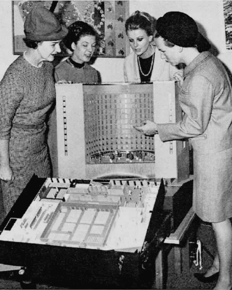 Four women in vintage clothing examine a large model of a building, possibly for architectural planning.