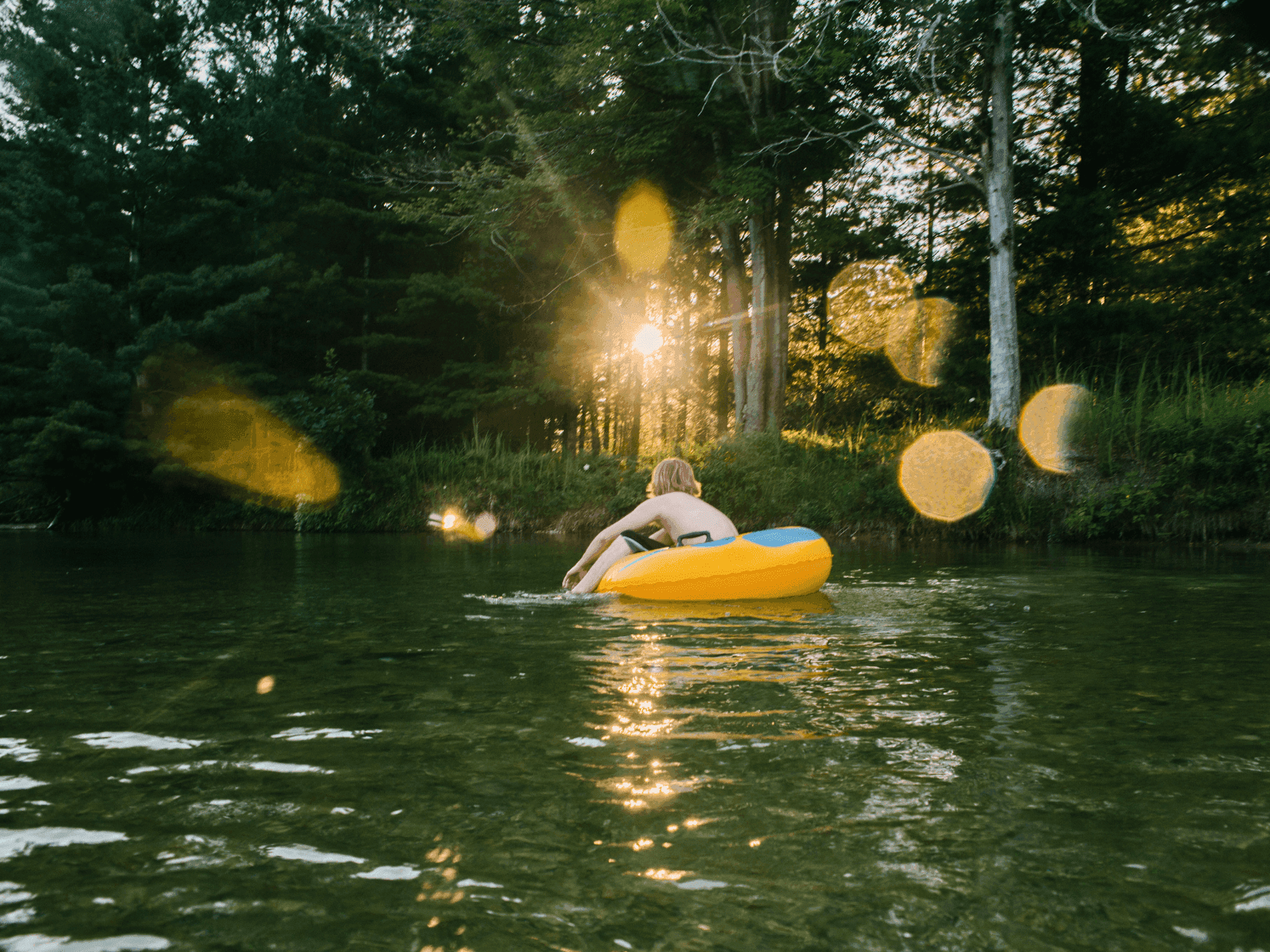 Man on yellow tube floating on river, trees in background, lens flare from sun.