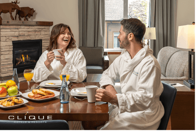 Cheerful couple in white robes enjoys breakfast at a wooden table in their Falcon Crest Lodge suite with a fireplace