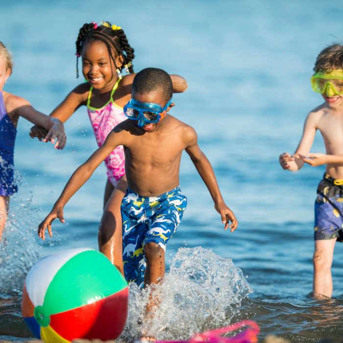 four young children in colorful bathing suits playing in the ocean with goggles and a beach ball