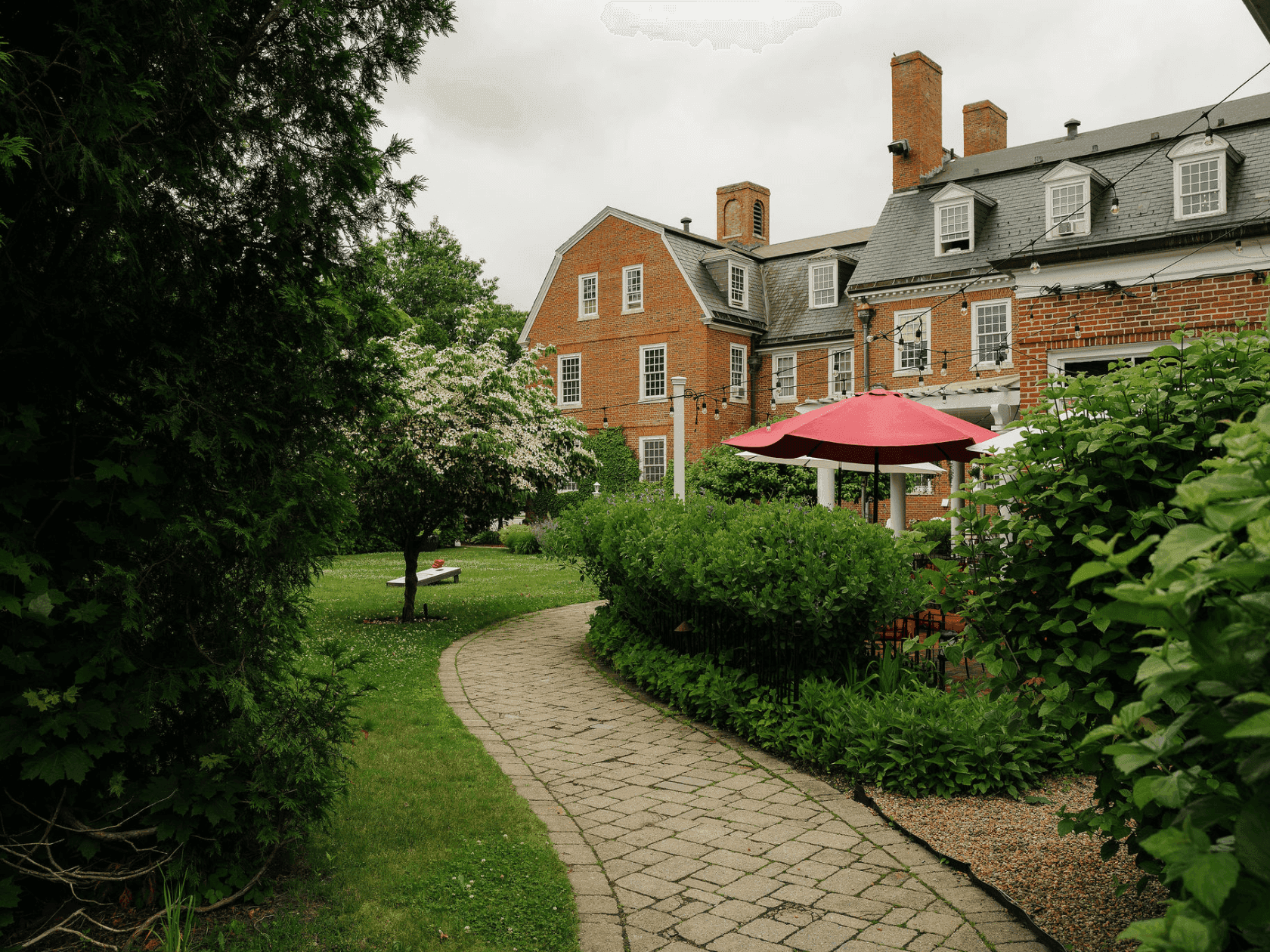 Brick path leads to a large brick building with two red umbrellas and a picnic table on the lawn.