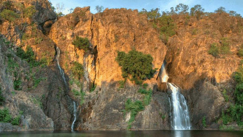 Waterfalls cascading down orange and gray rock cliffs surrounded by lush green vegetation under a clear blue sky.