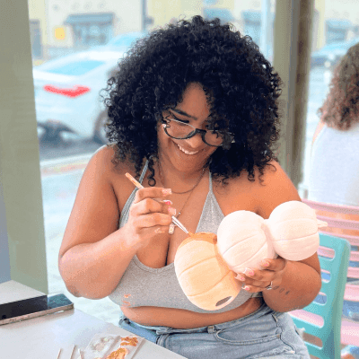 A curly-haired woman with glasses in jeans and a crop top sits and paints a stack of pumpkins in front of a bright window. All Fired Up is a great spot for DIY art in Orlando.