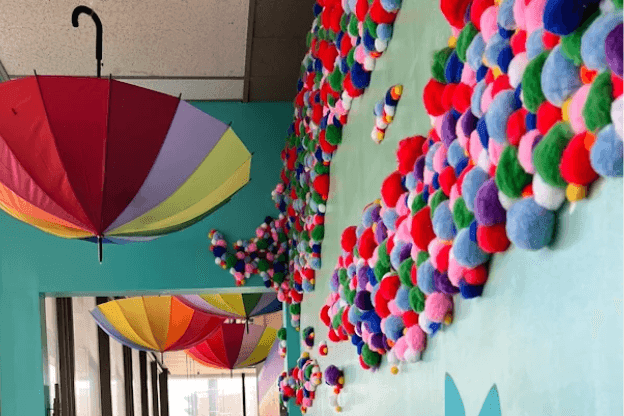 Photograph of a colorful indoor art installation featuring multiple upside-down umbrellas in rainbow colors suspended from a ceiling. The floor is decorated with numerous multicolored pom-poms arranged in swirling patterns against a light blue background, creating a vibrant and playful atmosphere.