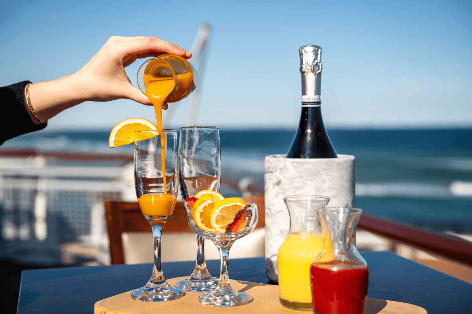 A hand pouring orange juice into glasses with champagne and a bottle on a table overlooking the ocean.
