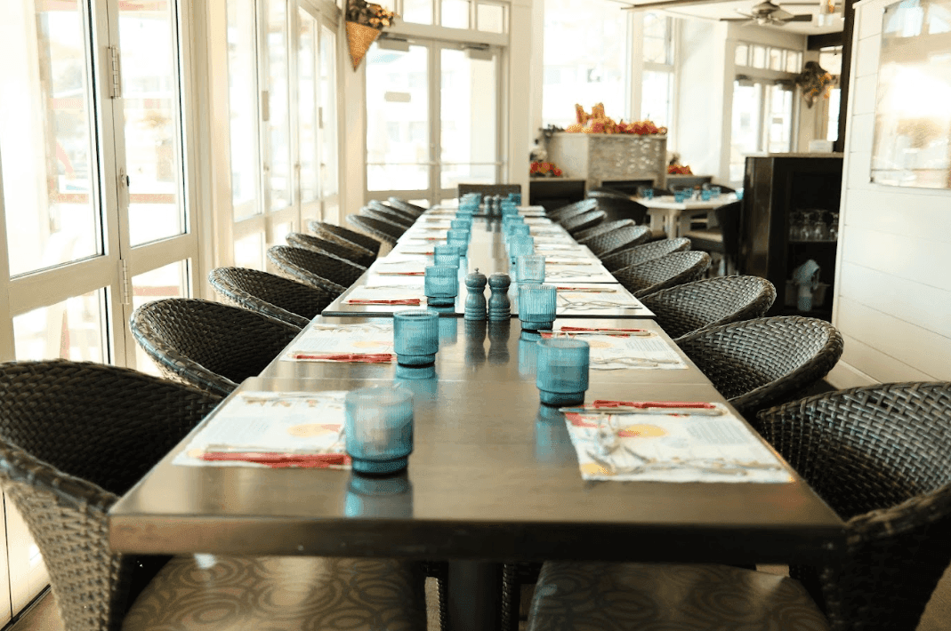 A long dining table set with blue glasses and wicker chairs in a bright room with glass doors.