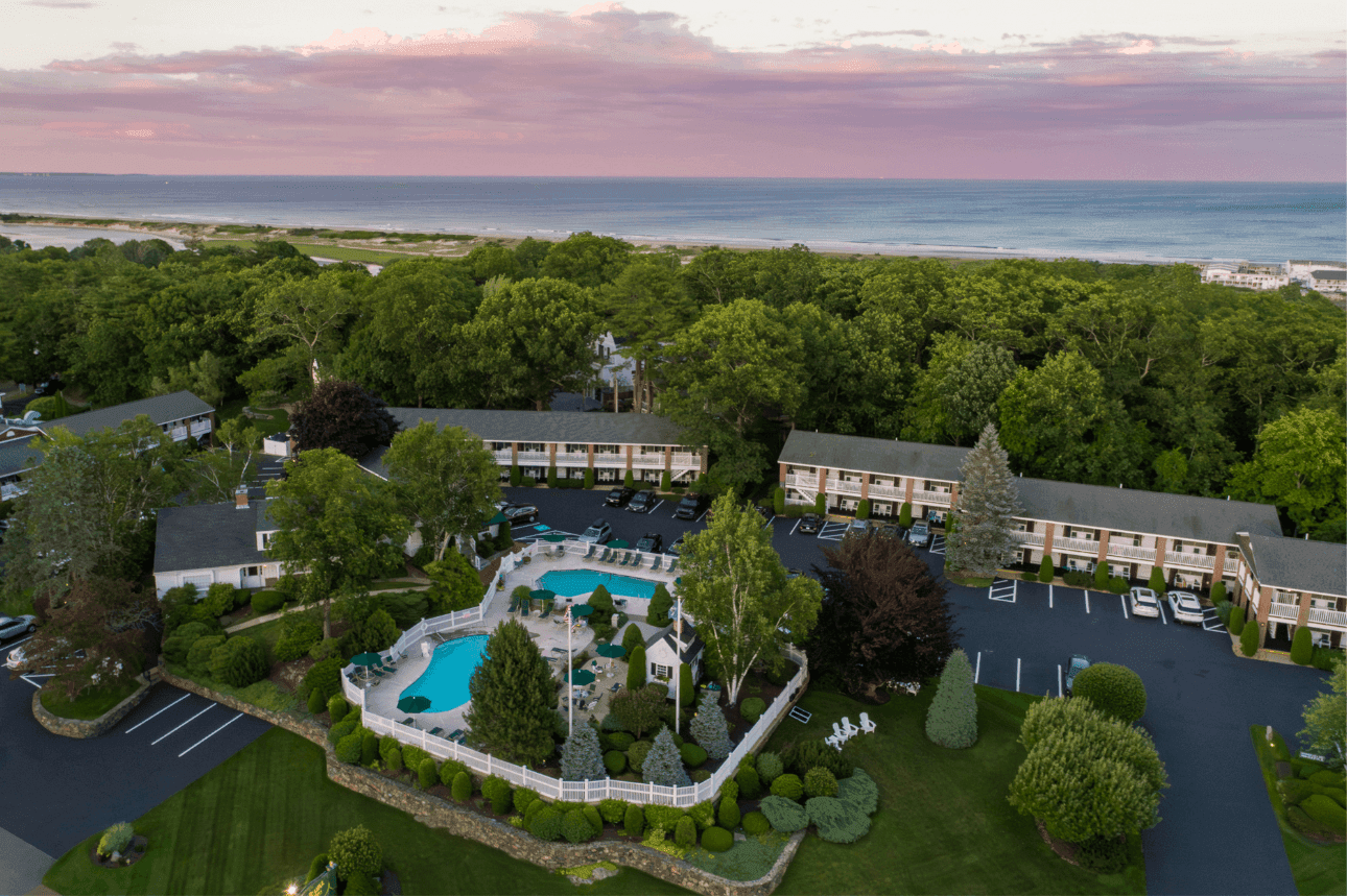 Aerial view of the Juniper Hill Inn with its pool and parking lot surrounded by trees and overlooking the ocean.