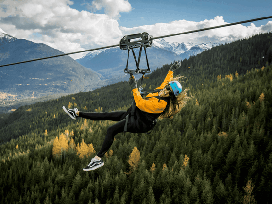 A woman wearing a helmet and harness rides a zipline over a forested area at Aava Whistler.