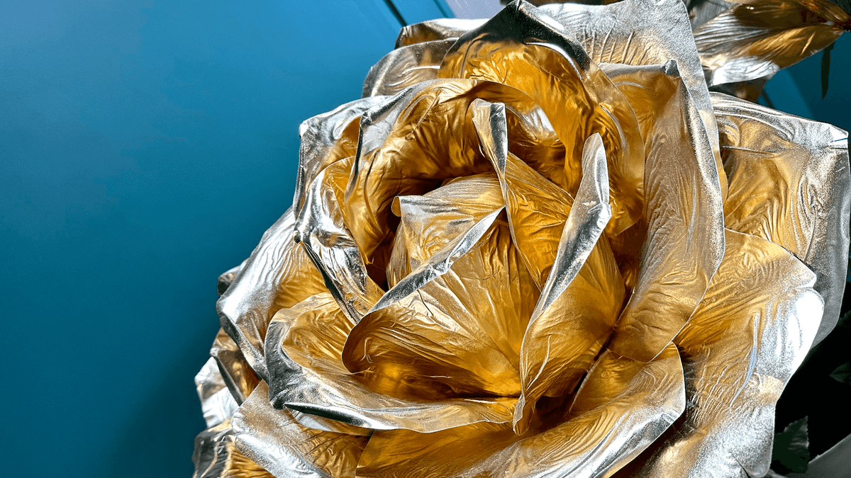 Rose en métal doré et argenté sur fond bleu clair, détaillant ses pétales brillants et textures.