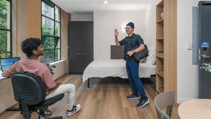 A man stands in a furnished room while another person sits nearby, both looking at each other.