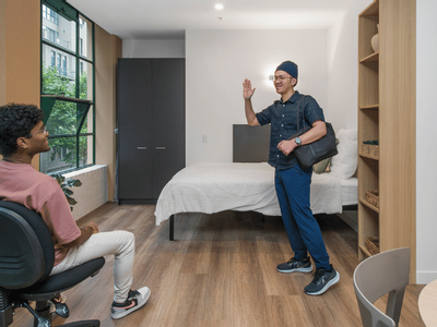 A man stands in a furnished room while another person sits nearby, both looking at each other.