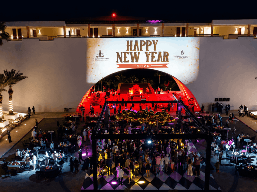 Aerial view of an elegant outdoor New Year's Eve celebration with illuminated stage, palm trees, and guests on a checkered floor.