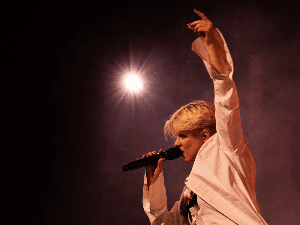 Blonde woman singing into microphone on stage under spotlight for Robyn's Sexistential Tour 2026.