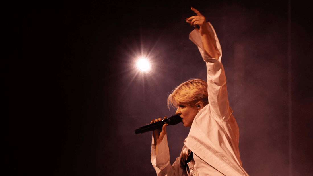 Blonde woman singing into microphone on stage under spotlight for Robyn's Sexistential Tour 2026.