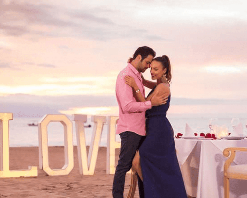 Couple hugging on the beach beside a glowing LOVE sign and a romantic dining table at Casa Dona Susana