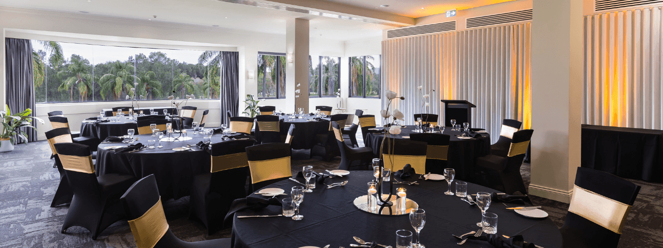 A large banquet hall with multiple round tables set for dining, featuring black tablecloths and yellow chairs.