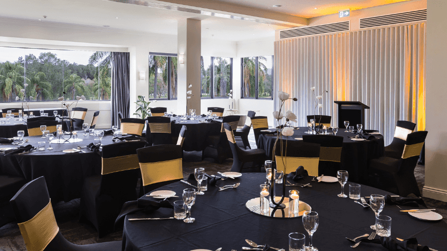 A large banquet hall with multiple round tables set for dining, featuring black tablecloths and yellow chairs.