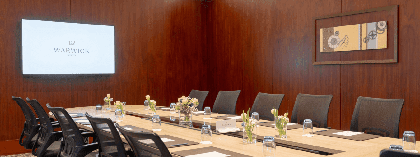 Gobelin with a long wooden table by black mesh chairs under a wall-mounted screen at Warwick Geneva