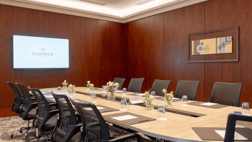 Gobelin with a long wooden table by black mesh chairs under a wall-mounted screen at Warwick Geneva