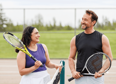 Couple holding tennis rackets and smiling together beside the outdoor tennis court at Rodd Crowbush Golf & Beach Resort