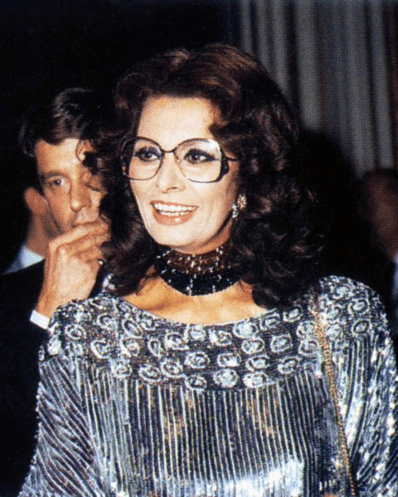 A smiling woman in glasses and a shiny dress, with a man behind her in a black suit.
