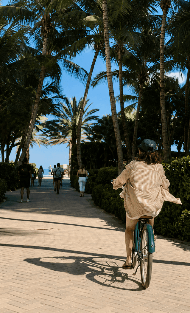 A woman rides a bicycle down a palm-lined pathway with other people walking in the distance.