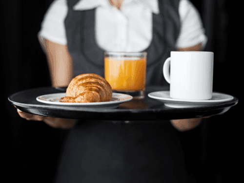 A waiter holds a tray with a fresh croissant, orange juice, and a coffee mug in The Croft House at The Sebel Brisbane