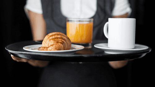 A waiter holds a tray with a fresh croissant, orange juice, and a coffee mug in The Croft House at The Sebel Brisbane