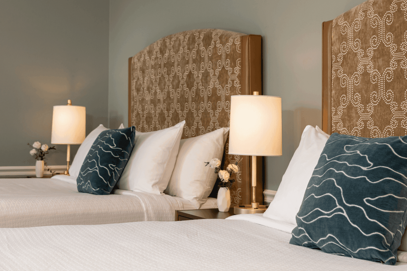 Two beds with patterned headboards and teal pillows by gold lamps and small flower vases at La Tourelle Inn and Spa
