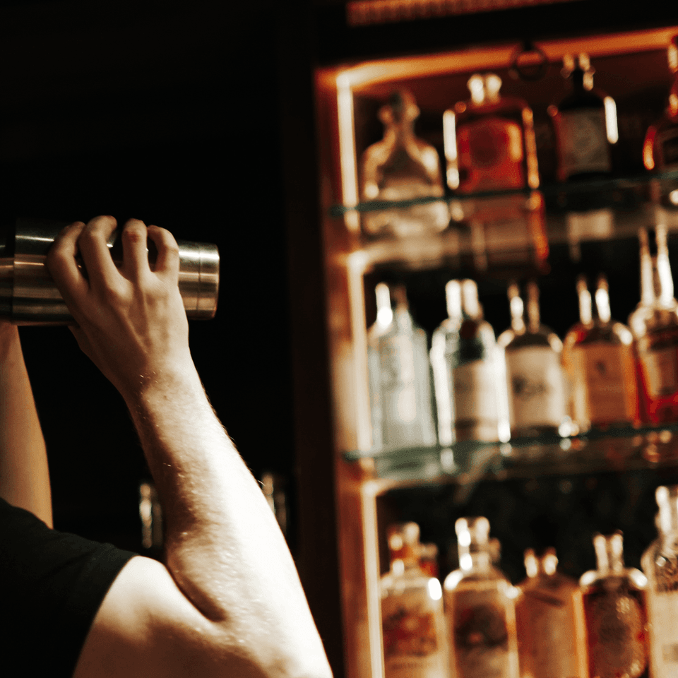 Bartender shaking a cocktail shaker beside a glowing cabinet of liquor bottles at the bar in La Tourelle Inn and Spa