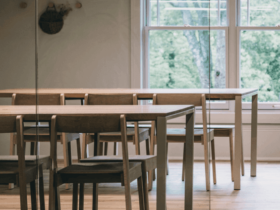 Wooden dining tables and chairs in a room with glass wall showing trees outside.