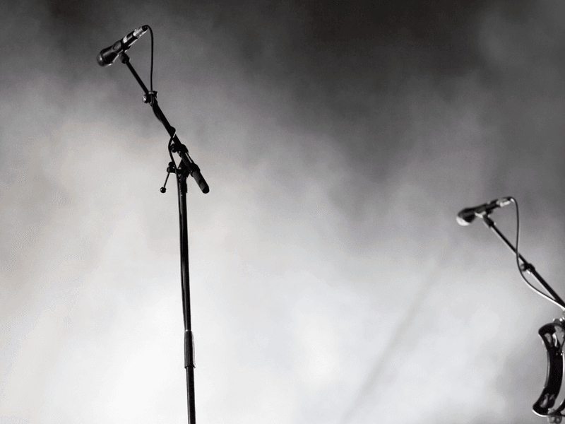 Two microphones on stands with a tambourine against a smoky black and white concert stage background.
