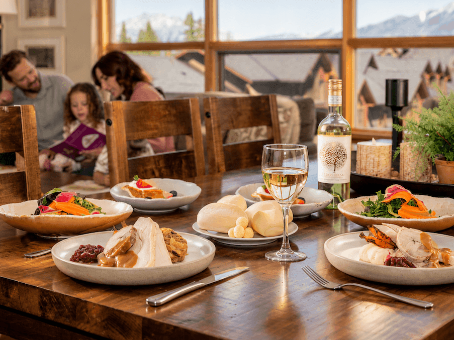 Family enjoying Thanksgiving Dinner To-Go with turkey, sides, wine, and rolls on a wooden dining table at home.