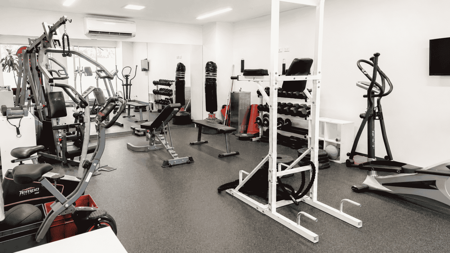 Modern fitness center with weight machines, free weights, cardio equipment, and exercise benches in a bright white room.