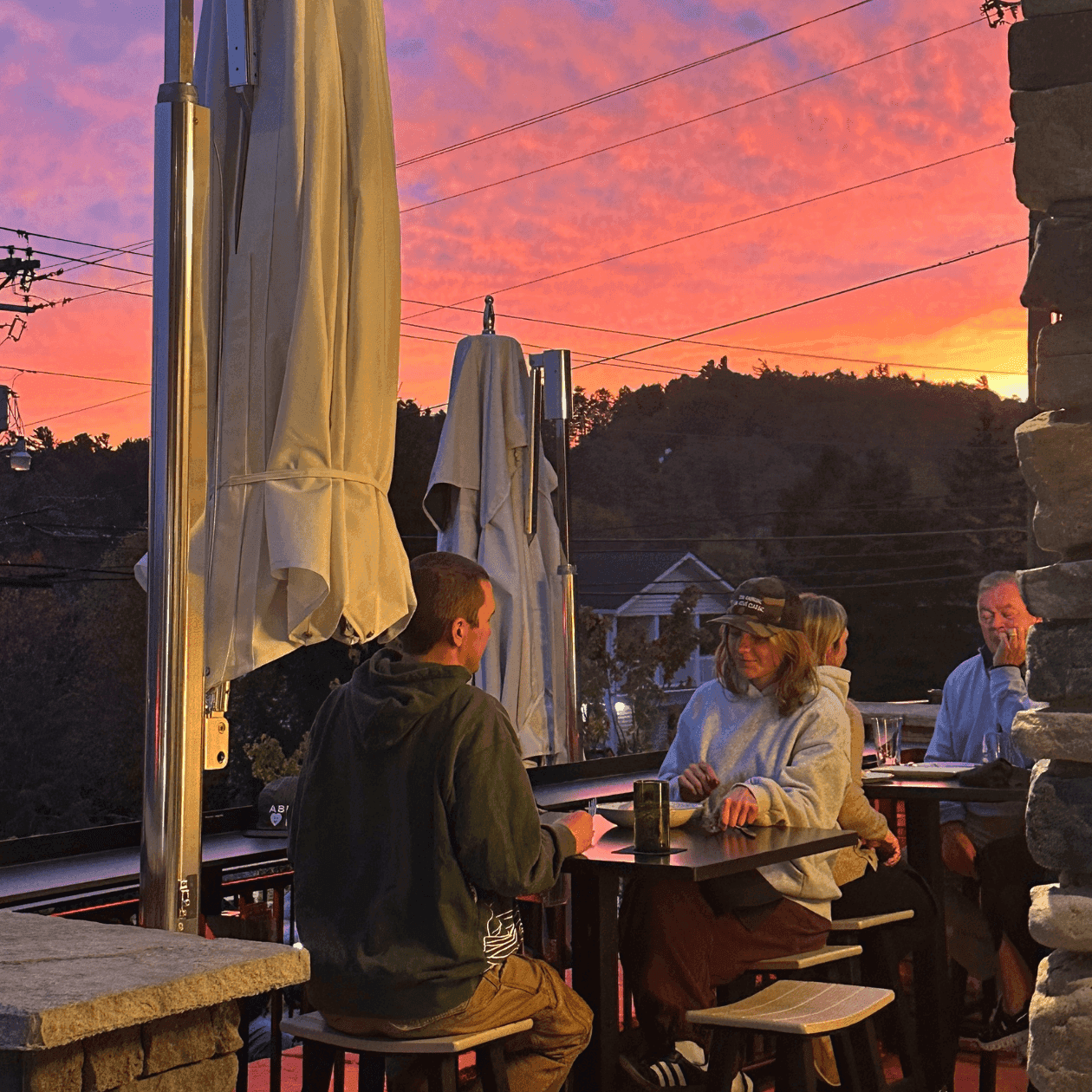People sitting at outdoor tables with umbrellas at dusk with a pink sky in the background.