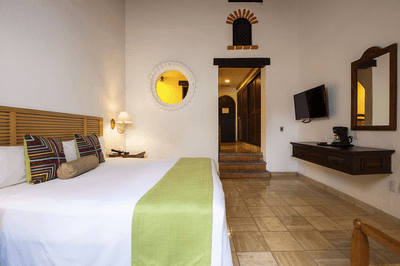Hotel room with tiled floor, bed, nightstand, wall-mounted lamp, TV, and mirror over a wooden shelf with a coffee maker.