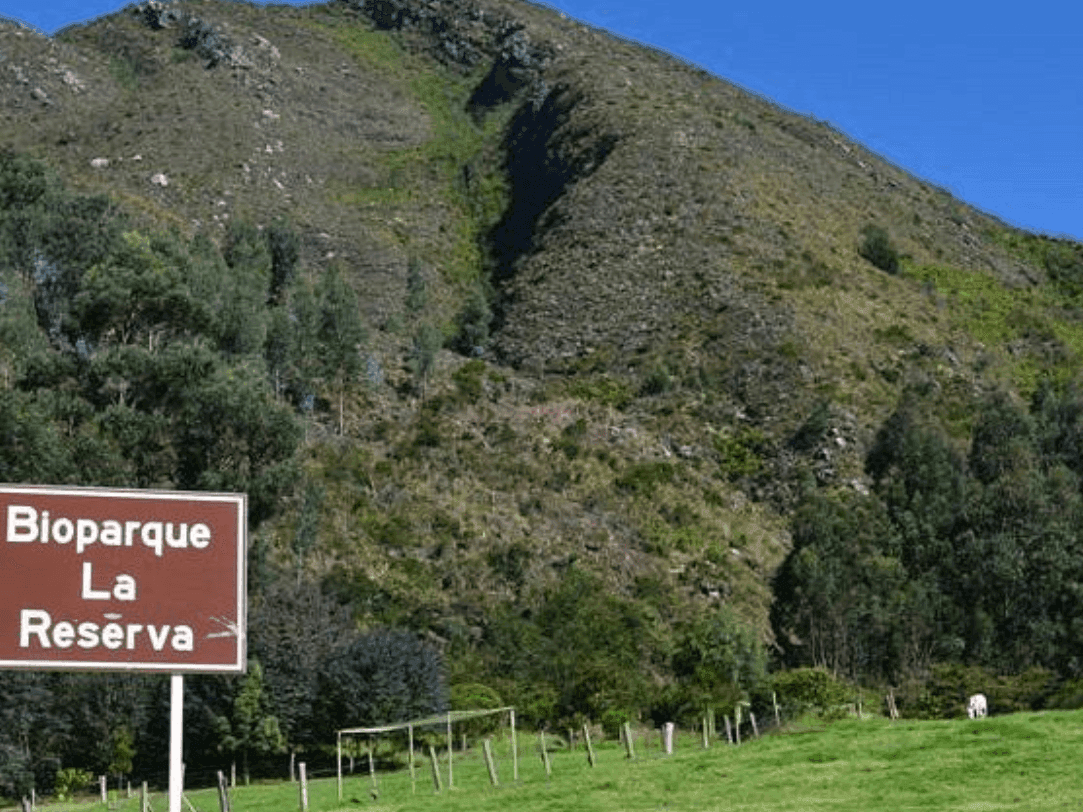 Cartel marrón con la inscripción 'Bioparque La Reserva' en una ladera cubierta de hierba con densos árboles verdes y un paisaje montañoso al fondo