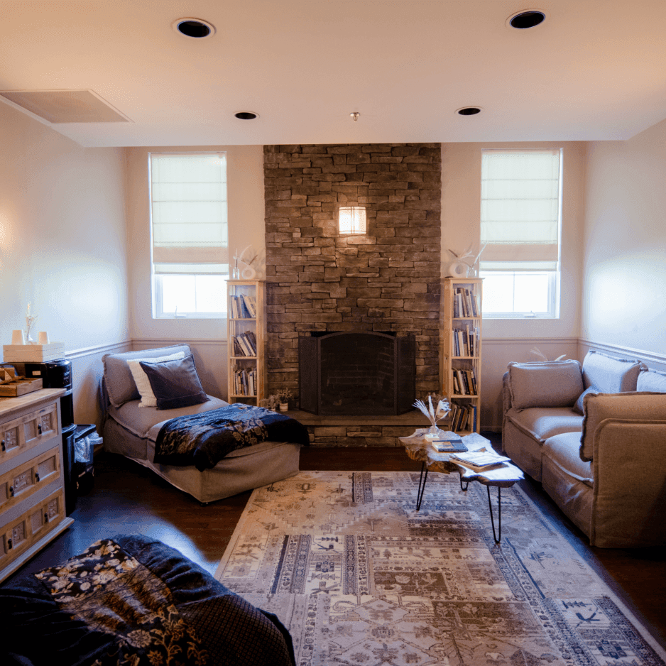 Spa lounge with gray chaise and sofas flanking a stone fireplace, bookshelves, and rug beneath at La Tourelle Inn & Spa