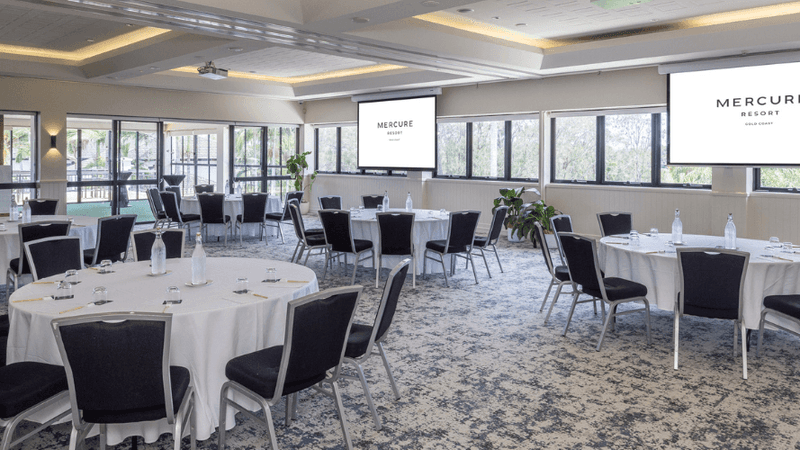 Banquet hall with round tables, black chairs, white tablecloths, two projectors, and large windows with outdoor view.