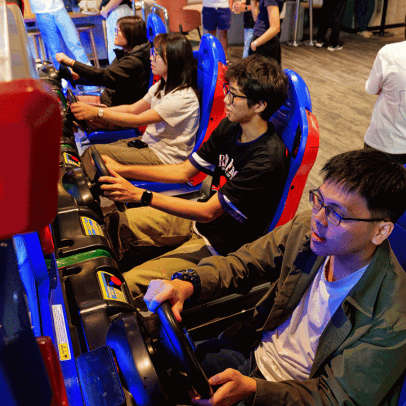 Students playing racing arcade games in a colorful gaming lounge with blue and red chairs at UniLodge.