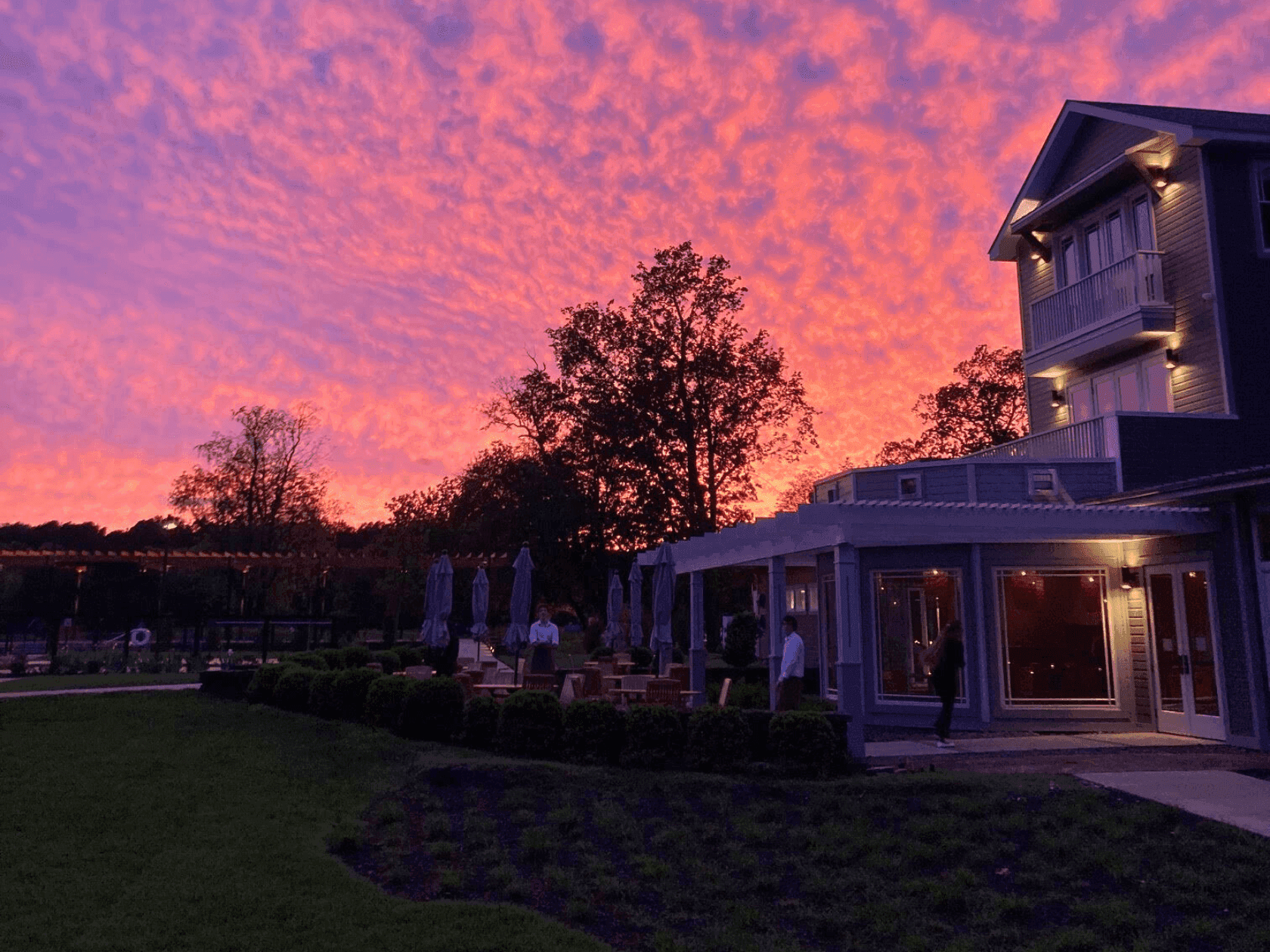 Pink sunset sky over a building with people and umbrellas on a patio.