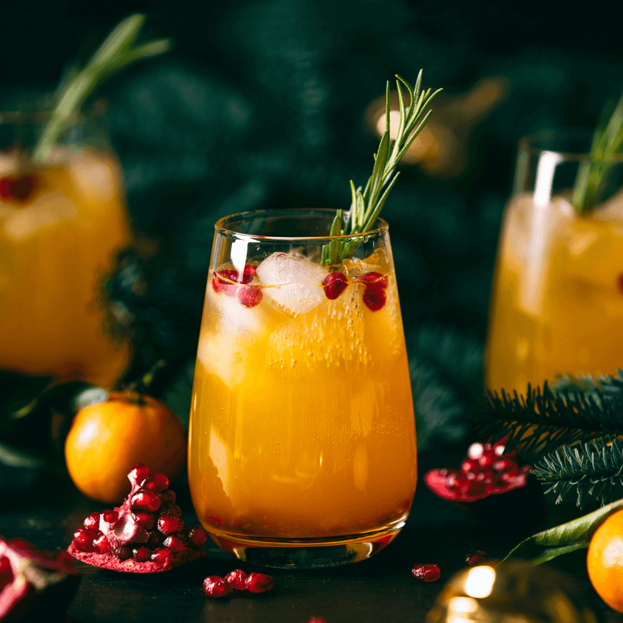 Three glasses of orange drink with rosemary and pomegranate seeds, surrounded by oranges and a Christmas tree branch.