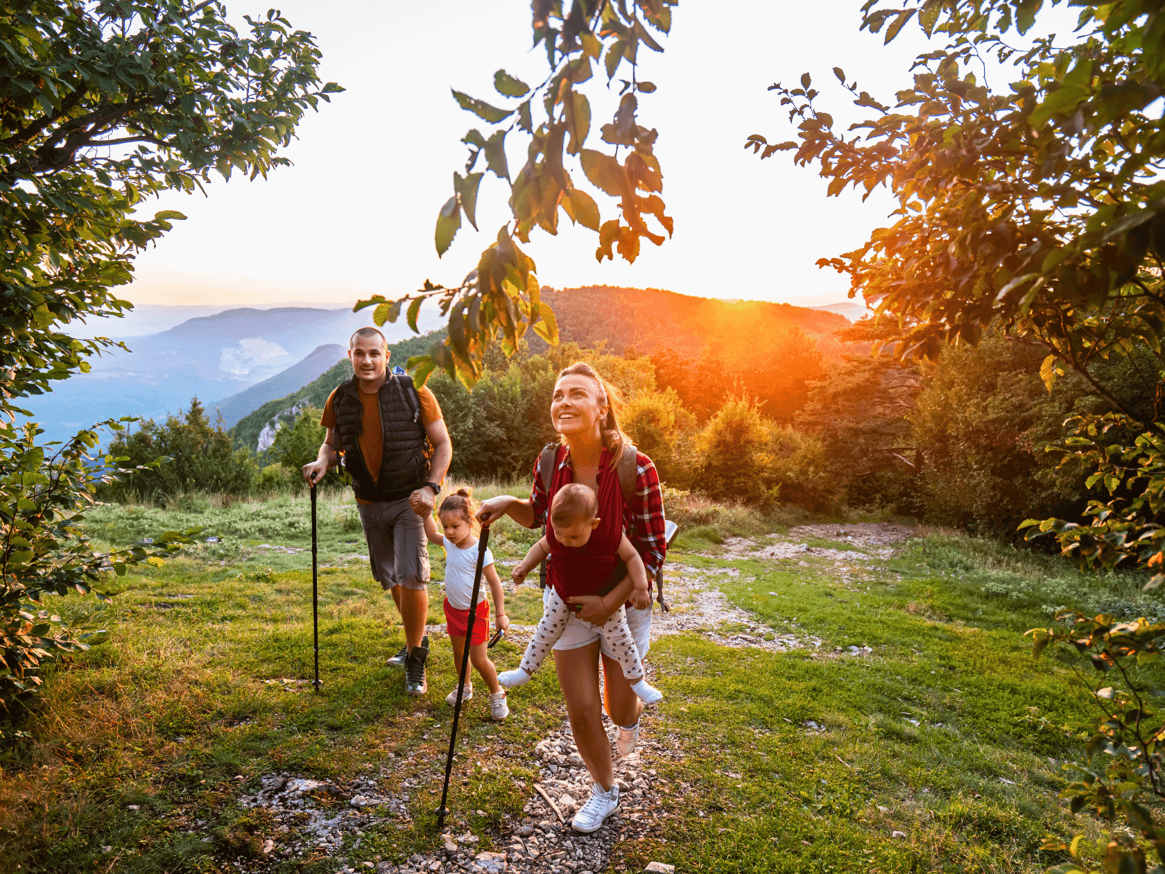 Family hiking in nature at sunset, man, woman, and two children with trekking poles, mountains in background.